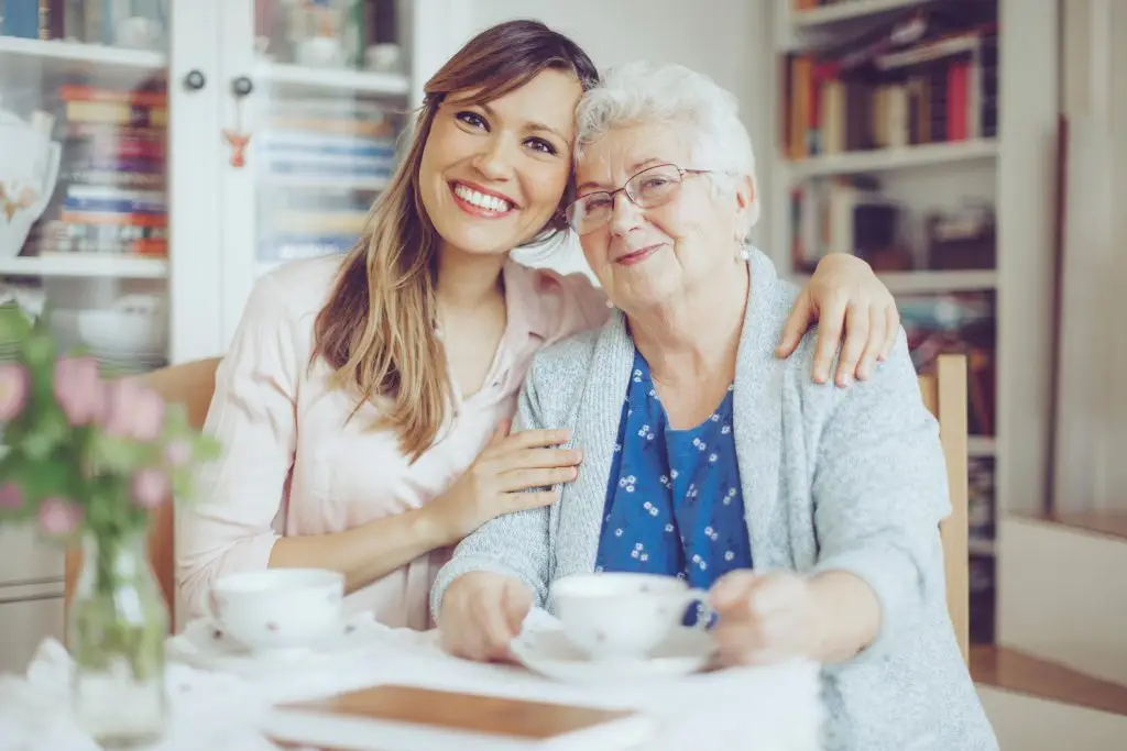 Social worker is visiting a senior woman in her own apartment. They are celebrating the elderly lady's birthday. The kind nurse is hugging the senior woman over her shoulder.