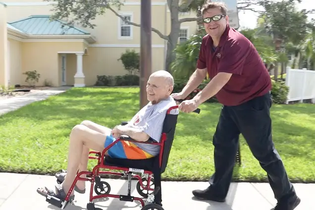 Smiling , male VNA hospice patient in wheelchair being pushed by a transport specialist.