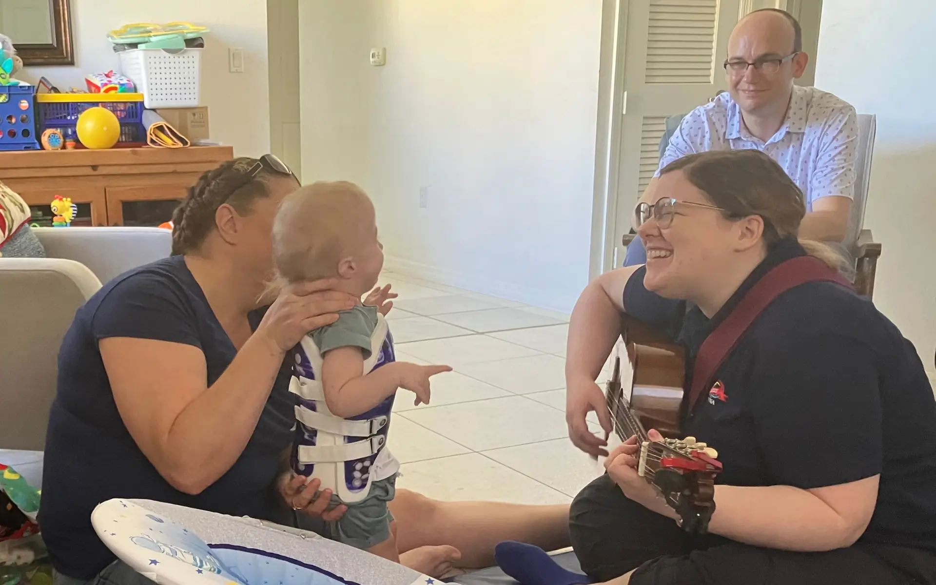 mother holding her baby as he listens to music therapist play guitar